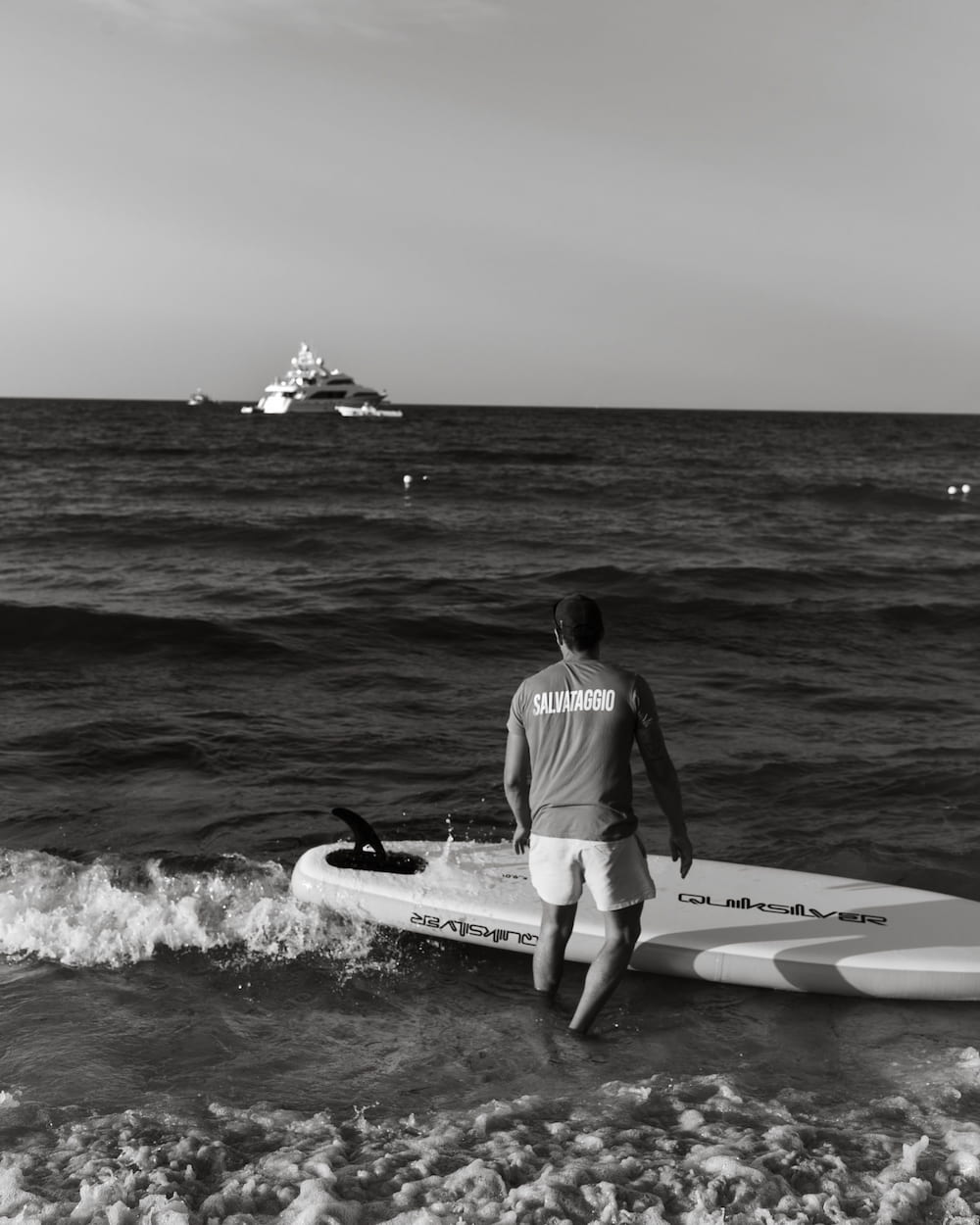 Black and white lifeguard in a "SALVATAGGIO" shirt pushing a paddleboard into the sea toward a distant yacht